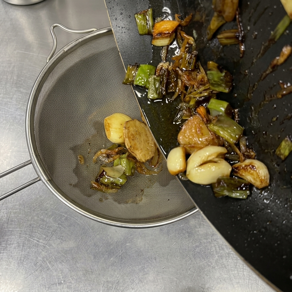 Hot seasoned oil and browned aromatics like garlic and scallions being poured from a dark pan into a fine-mesh strainer.