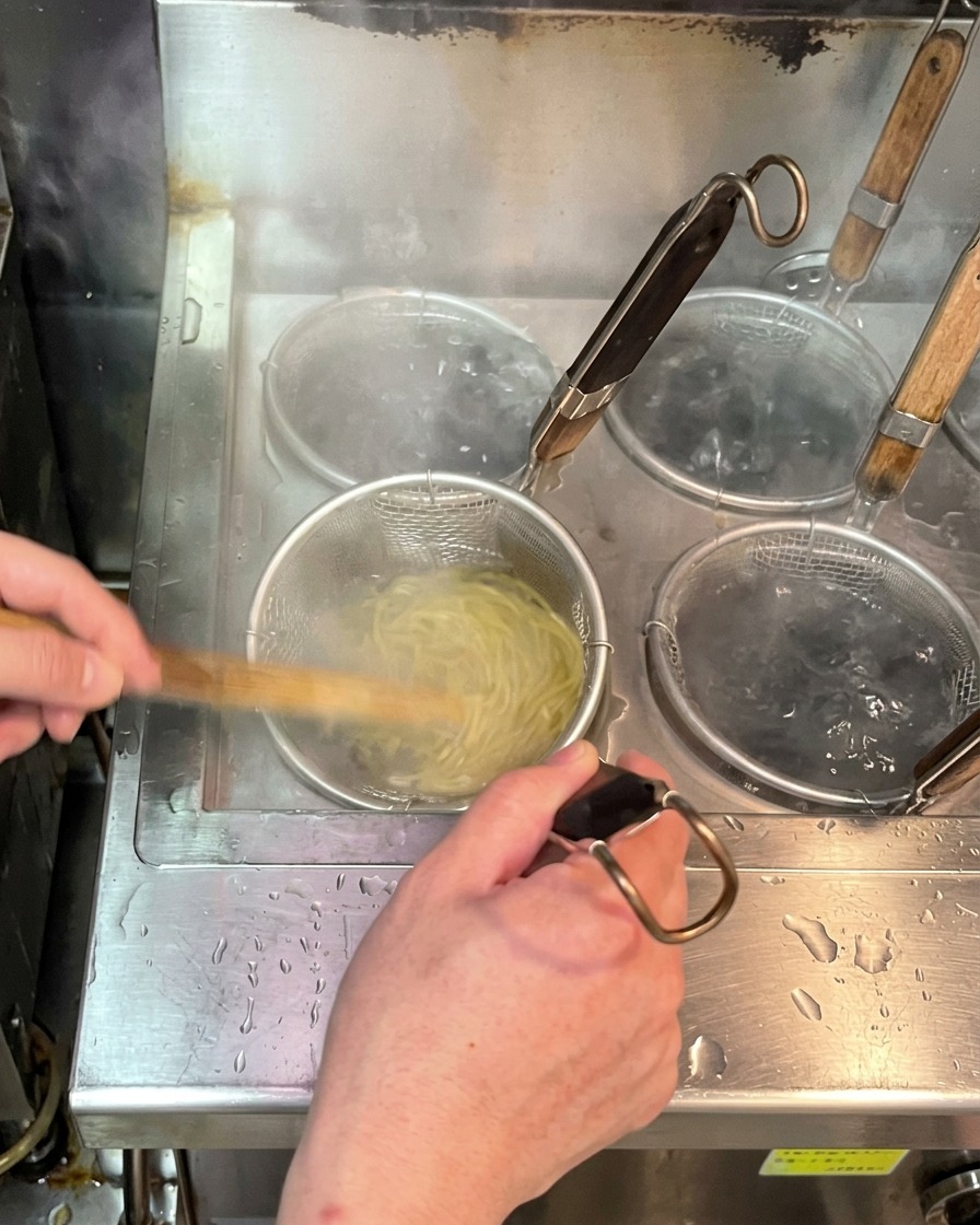Fresh ramen noodles cooking inside a submerged wire noodle basket in a pot of vigorously boiling water.