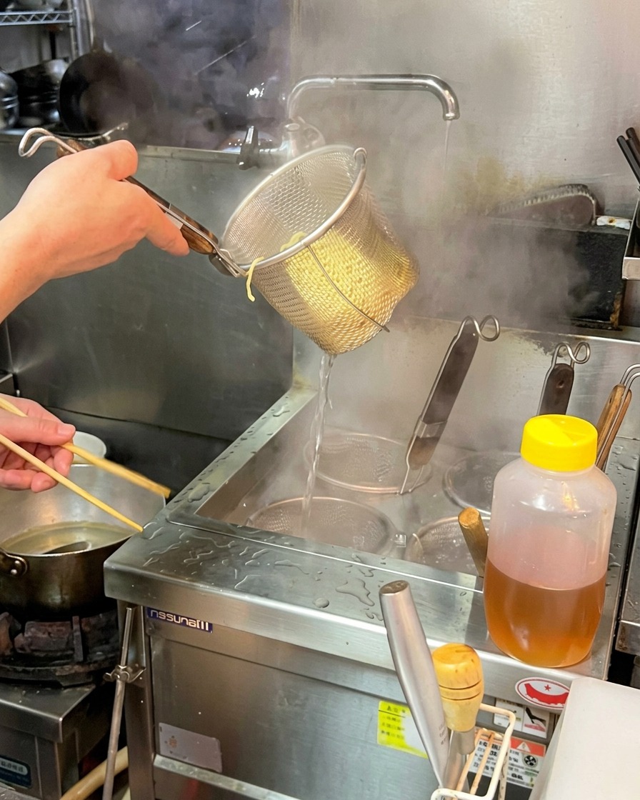 A chef firmly shaking a wire basket full of cooked ramen noodles to drain off excess hot water.