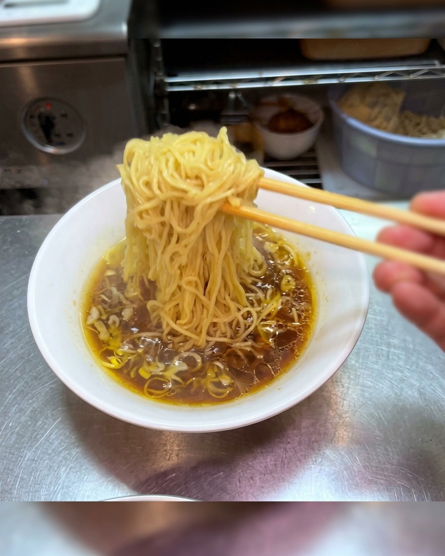 Chopsticks lifting a bundle of fresh ramen noodles high above a bowl of dark shoyu broth.
