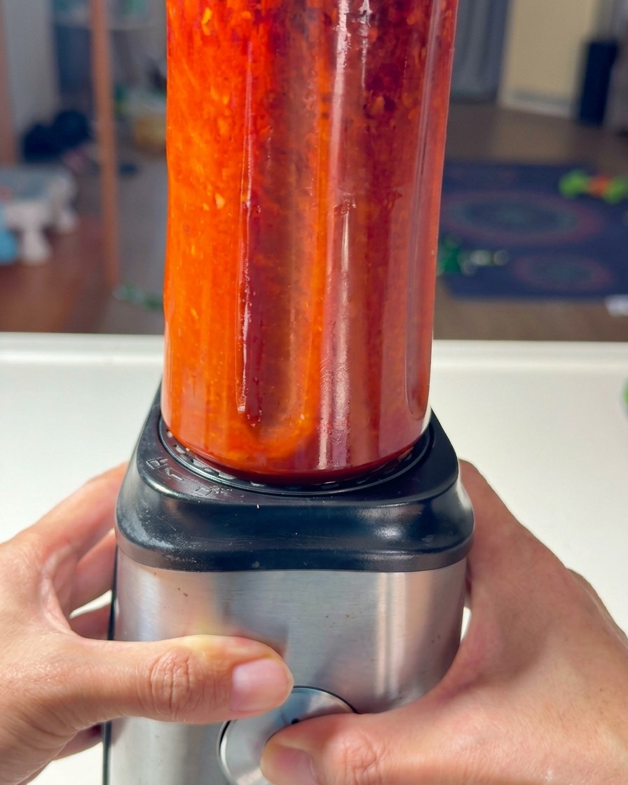 Two hands securing a tall blender cup filled with vibrant red chili paste onto a metal blender base.
