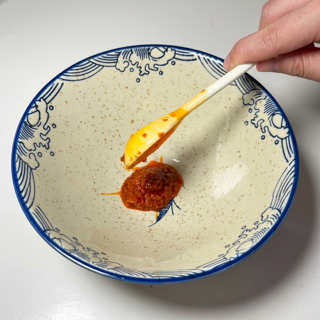 A spoonful of bright red chili paste being placed into an empty ceramic bowl.