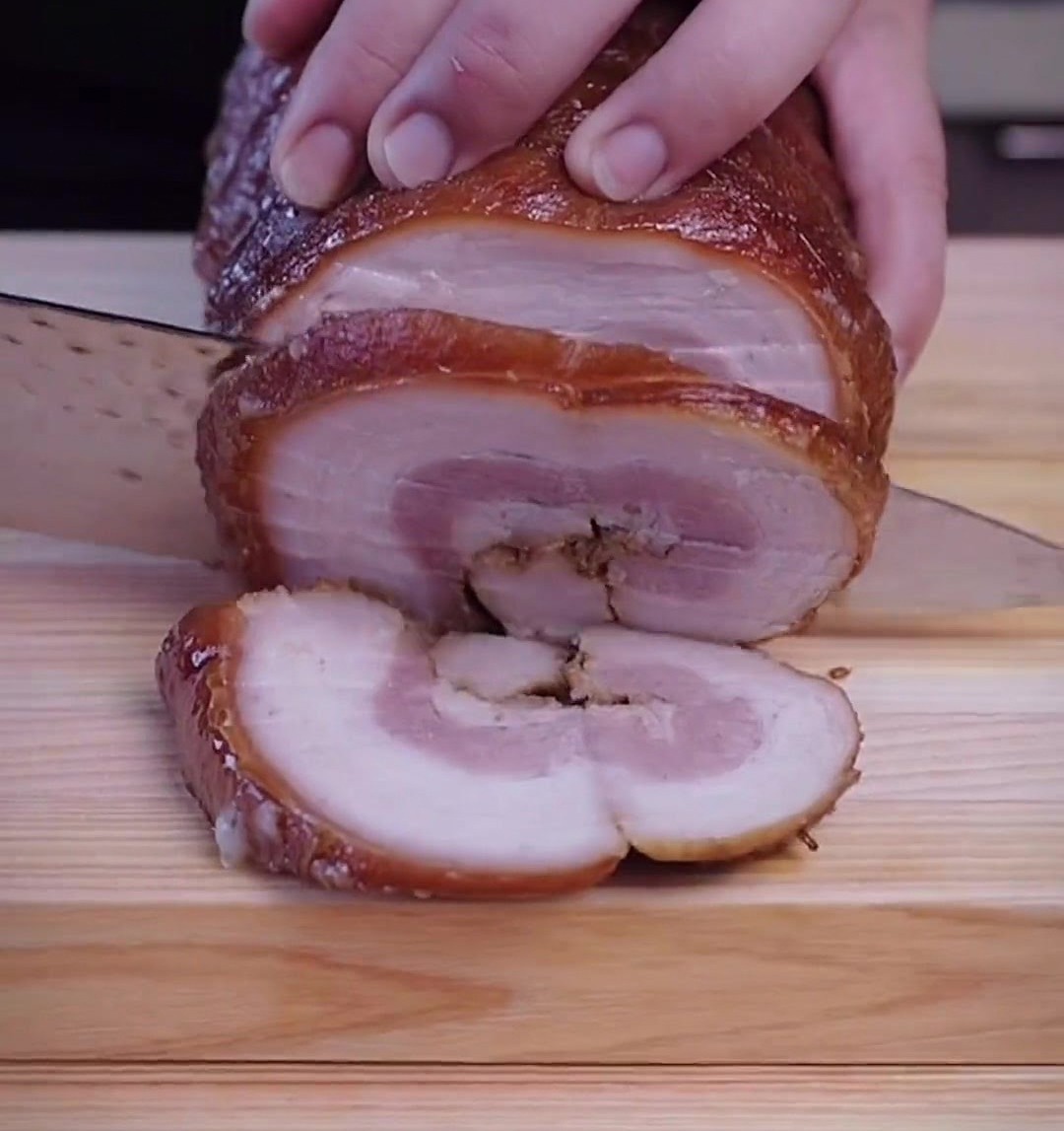 Hands holding a sharp knife, carefully slicing a browned, chilled chashu pork roll into thick rounds on a wooden cutting board.