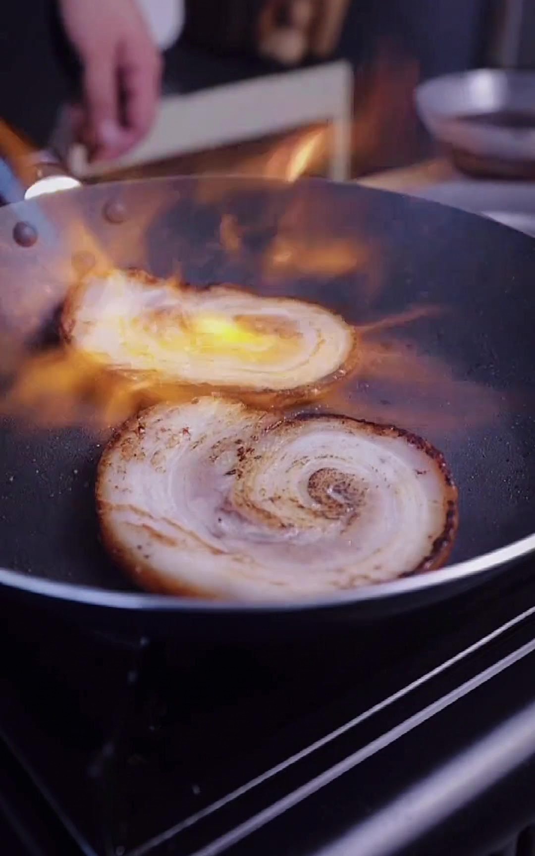 Two slices of chashu pork in a dark frying pan being hit with a bright orange flame from a kitchen blowtorch.