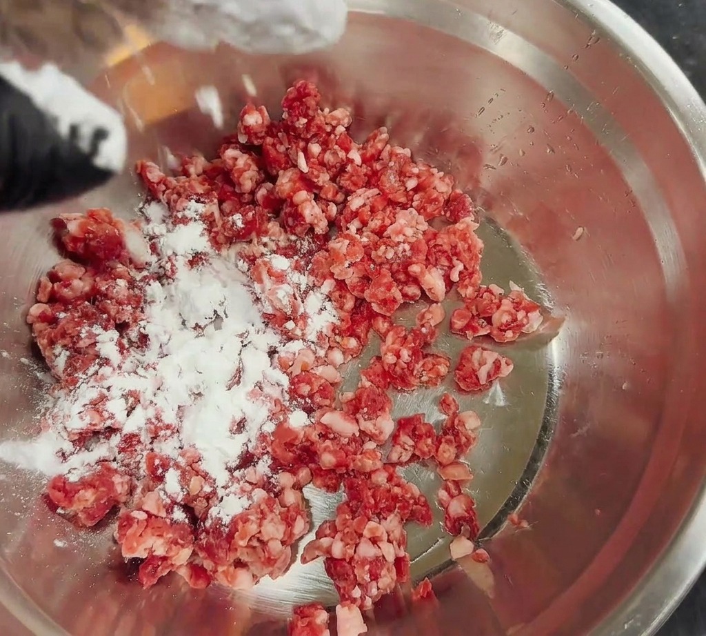 White cornstarch being dusted over a pile of fresh ground beef in a stainless steel bowl.