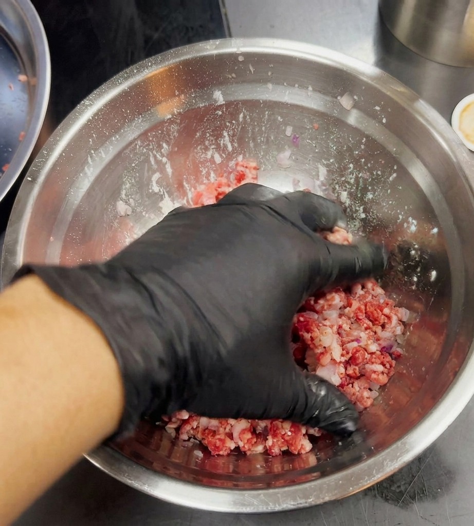 A black-gloved hand lightly mixing ground meat and minced purple onions in a silver bowl.
