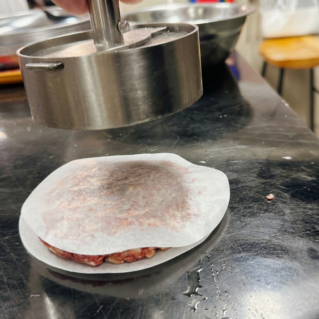 A cylindrical metal burger press shaping ground beef into a patty over a piece of parchment paper.