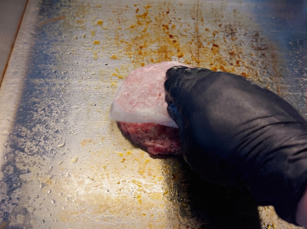 A gloved hand peeling parchment paper off a perfectly round, raw beef patty on a hot metal griddle.