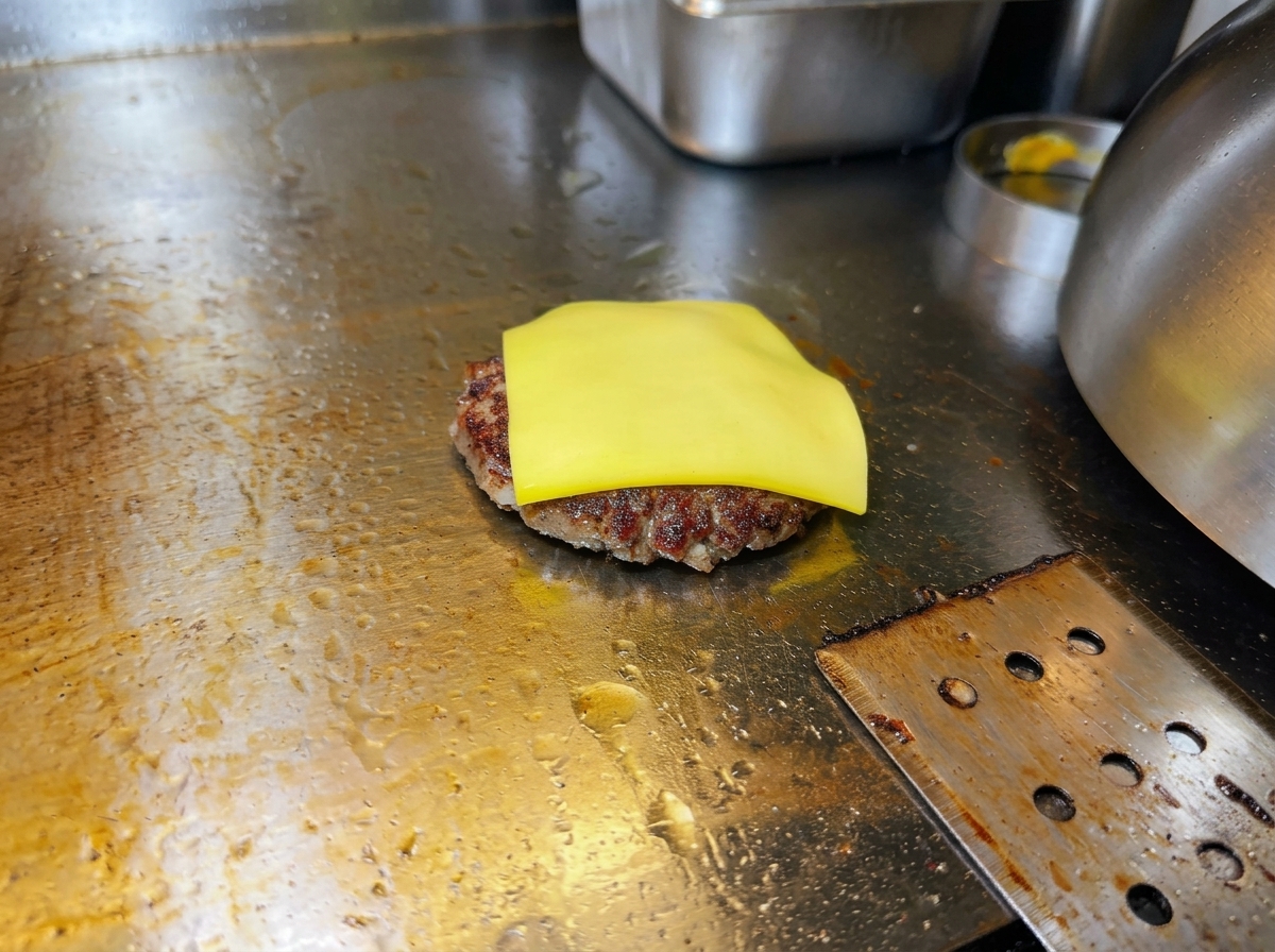 A slice of yellow cheese beginning to melt on top of a seared beef patty on a hot griddle.