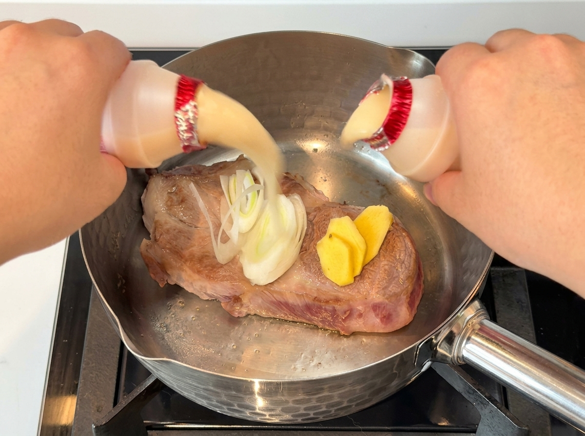 Hands pouring two small bottles of yogurt drink over a piece of seared pork with onions and ginger in a stainless steel pot.