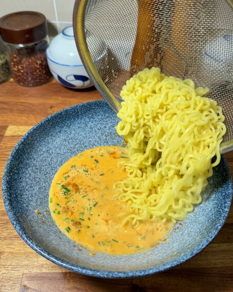 Cooked, wavy yellow ramen noodles sliding out of a metal strainer into a bowl of rich orange ramen broth.