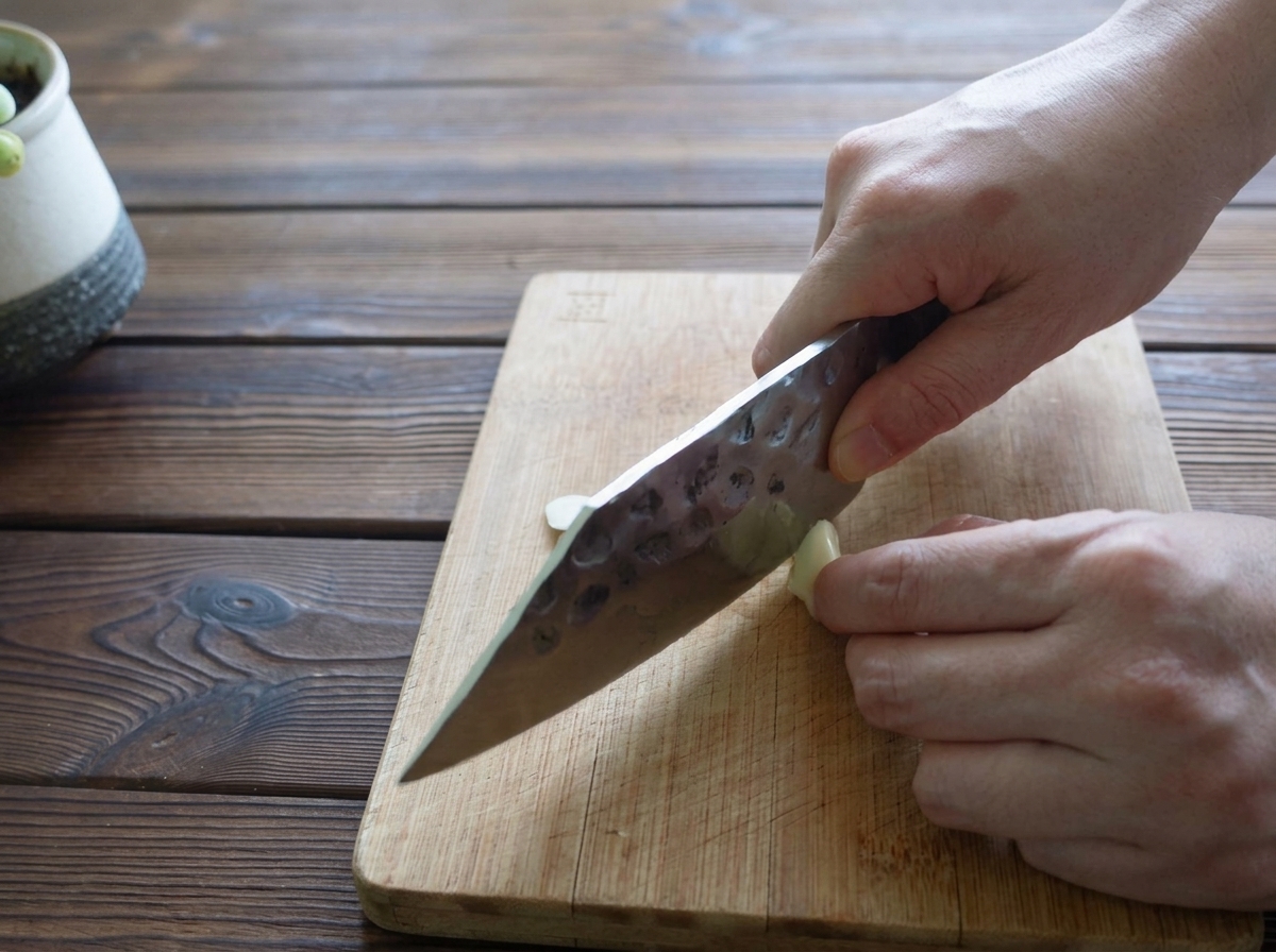 A persons hands using a knife to thinly slice a single clove of garlic on a wooden cutting board.