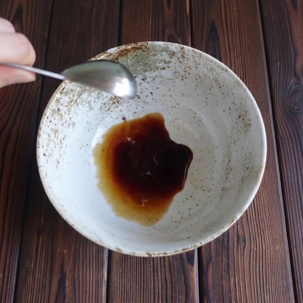 A measuring spoon pouring dark soy sauce into a speckled ceramic serving bowl.