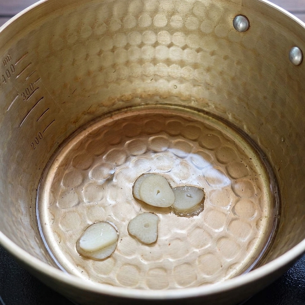 Thin slices of garlic frying gently in a pool of hot, clear melted pork lard inside a metal pot.