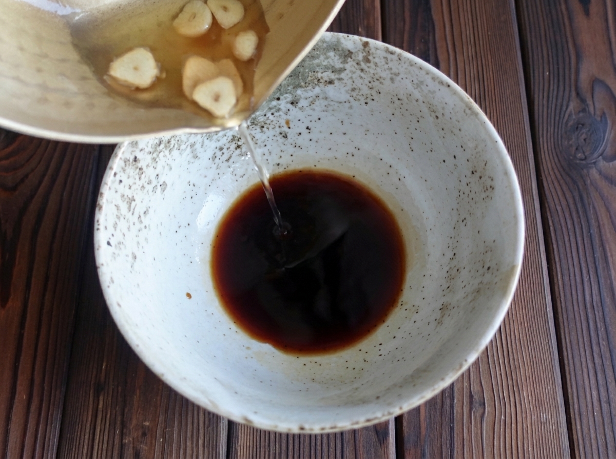 Hot garlic oil and toasted garlic slices being poured from a pan into a speckled bowl of dark soy sauce.