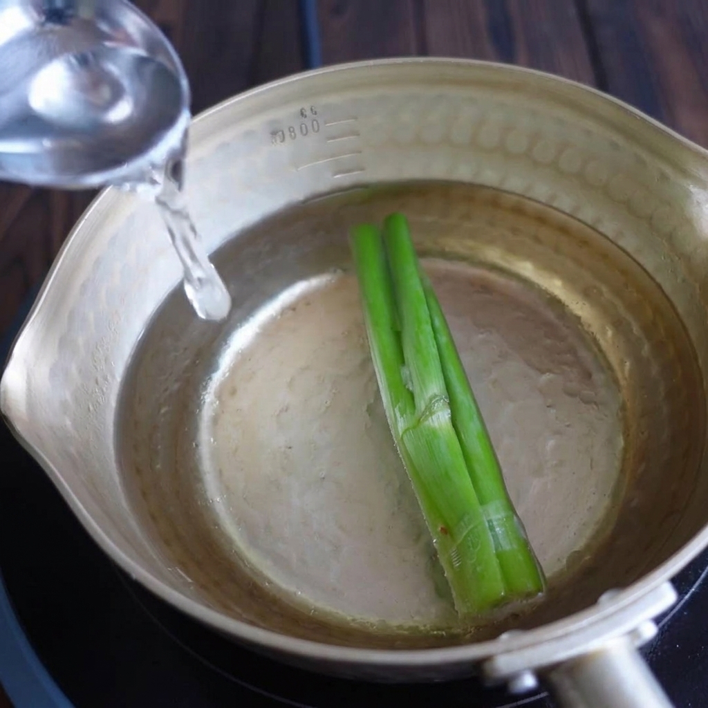 Pouring clear water from a vessel into a brass pot containing green scallion stalks.