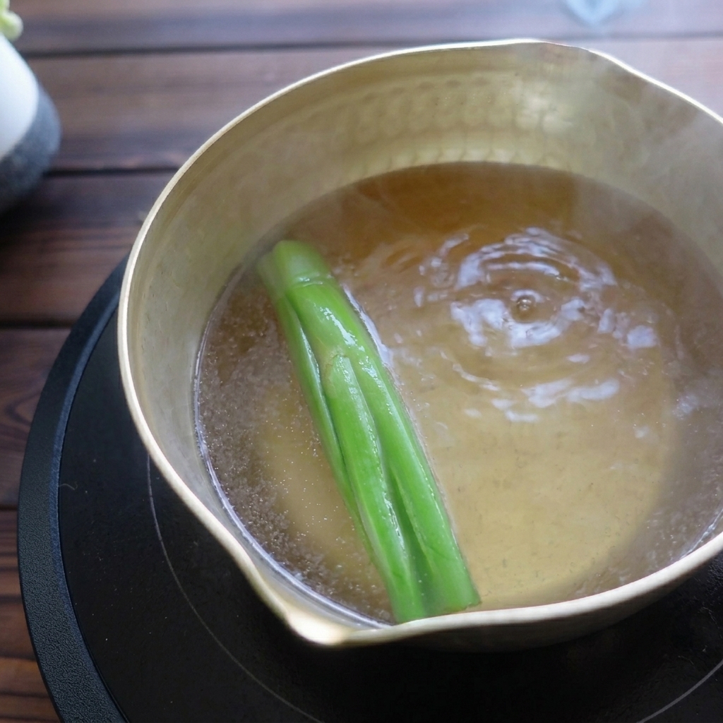 A brass pot simmering clear broth and green scallion stalks on a black stovetop.