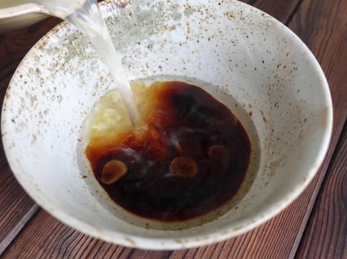 Pouring clear hot broth from a pot into a rustic ceramic bowl containing a dark, rich soy sauce mixture.