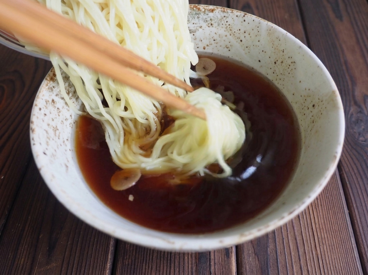 Wooden chopsticks gently lowering cooked ramen noodles into a bowl of dark, rich soy sauce broth.