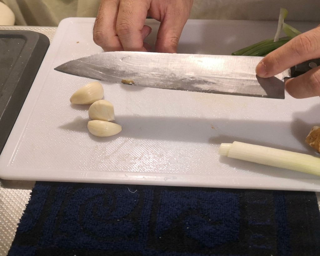 A person pressing the flat blade of a large kitchen knife over peeled garlic cloves to crush them on a cutting board.