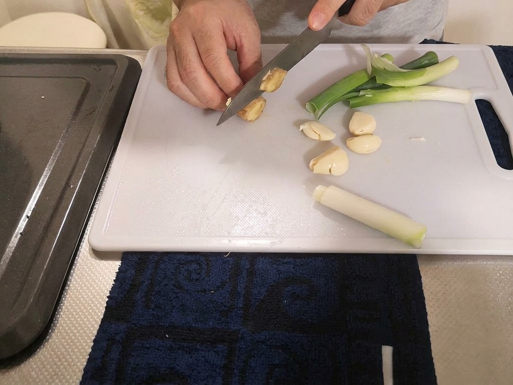 A cook slicing a piece of fresh ginger into thick pieces on a cutting board with scallions and garlic nearby.