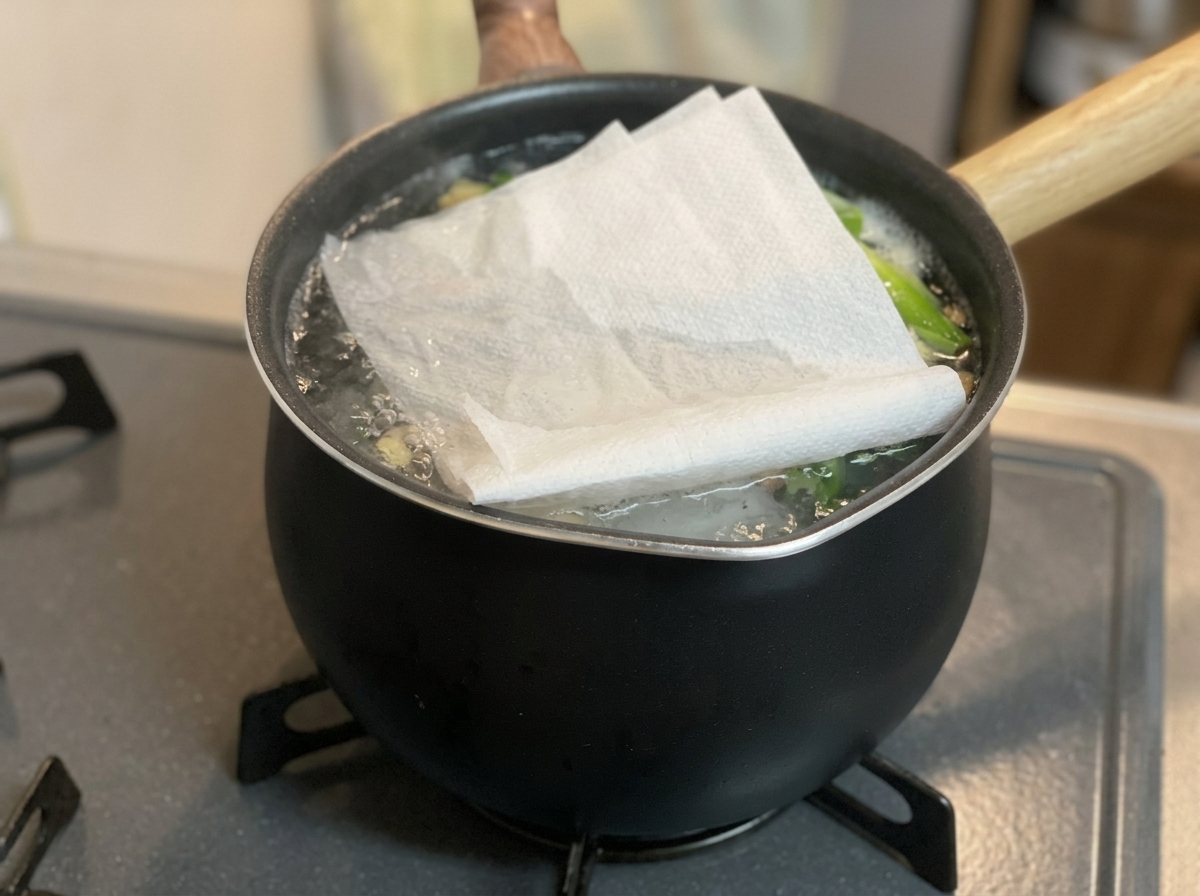 A folded white paper towel placed over the surface of a bubbling broth in a black pot on a stovetop.