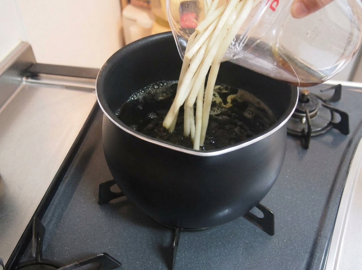 Pouring liquid and aromatics from a clear measuring cup into a simmering pot of dark broth.