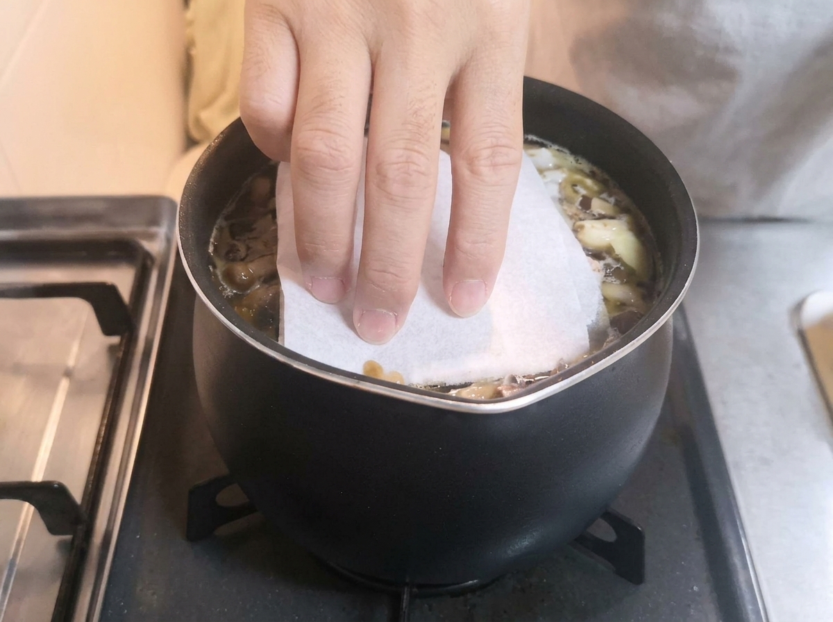 A hand placing a white paper towel over a simmering pot of dark broth and aromatics.