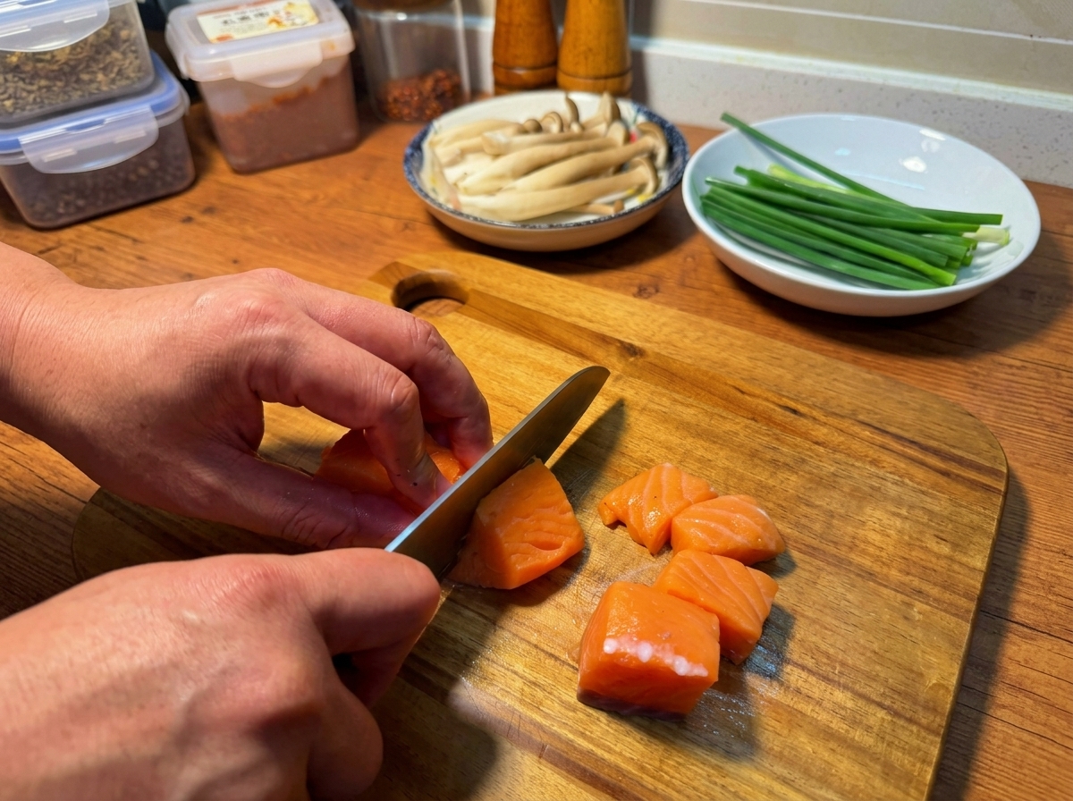 Hands using a knife to slice a fresh raw salmon fillet into even chunks on a wooden cutting board.