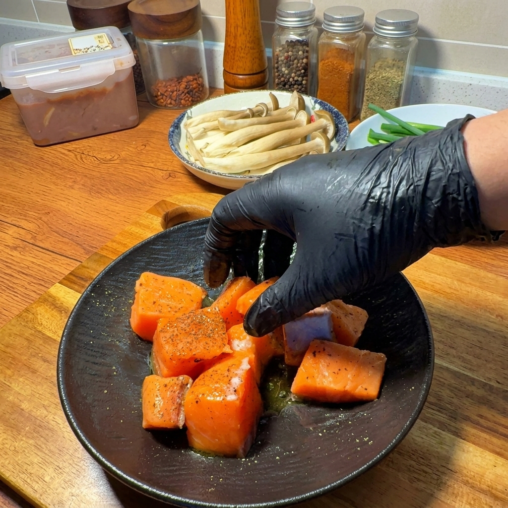 A gloved hand tossing raw salmon chunks with seasoning and olive oil on a dark plate.