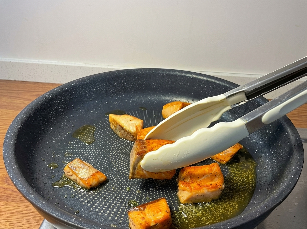 Tongs flipping golden-brown pan-fried salmon pieces in a non-stick skillet.