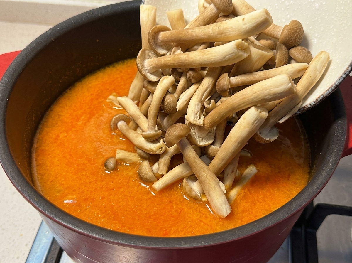 Pouring fresh brown beech mushrooms from a bowl into a pot of simmering orange spicy miso broth.