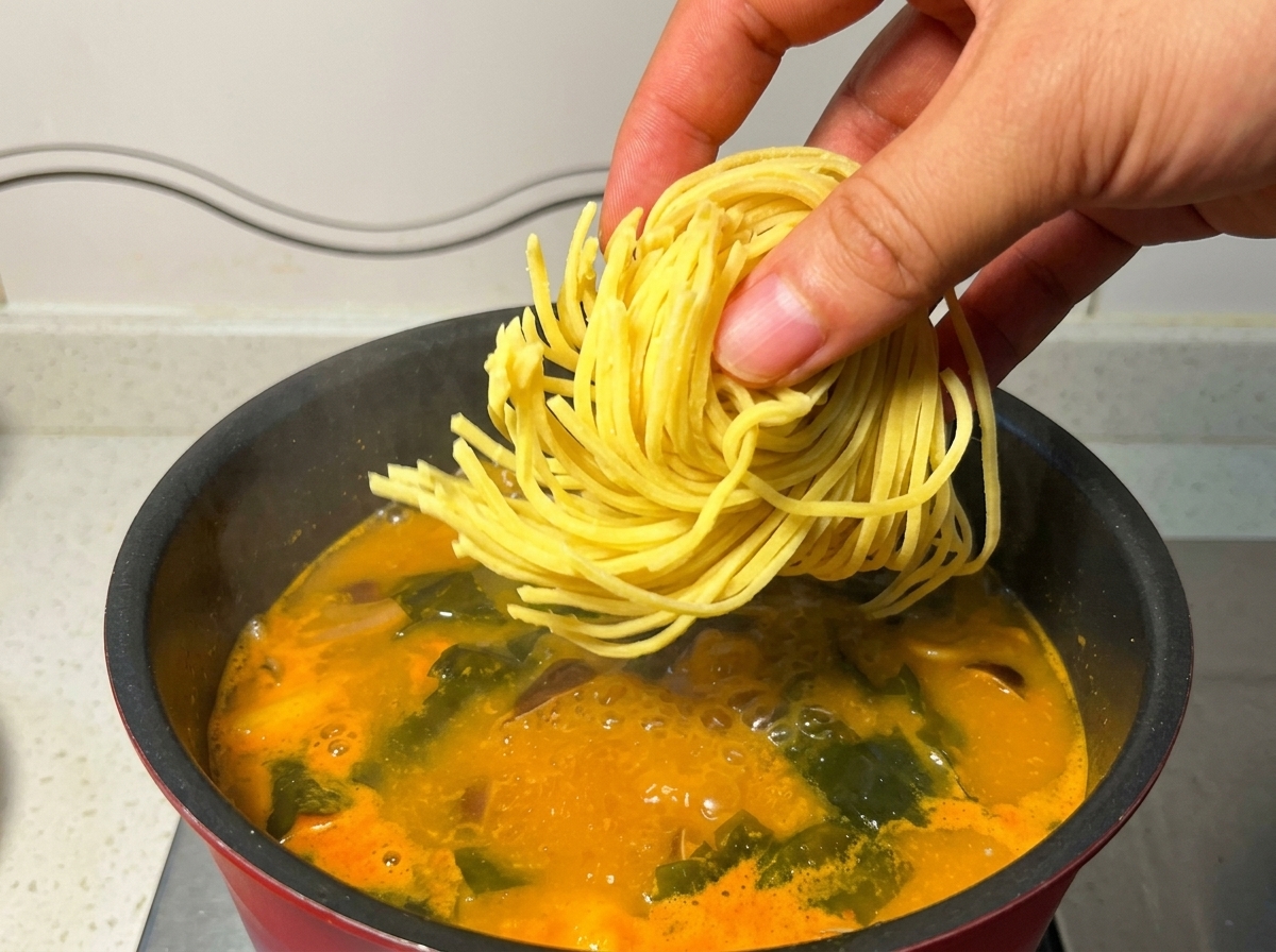 A hand dropping a nest of fresh yellow ramen noodles into a pot of boiling orange soup.