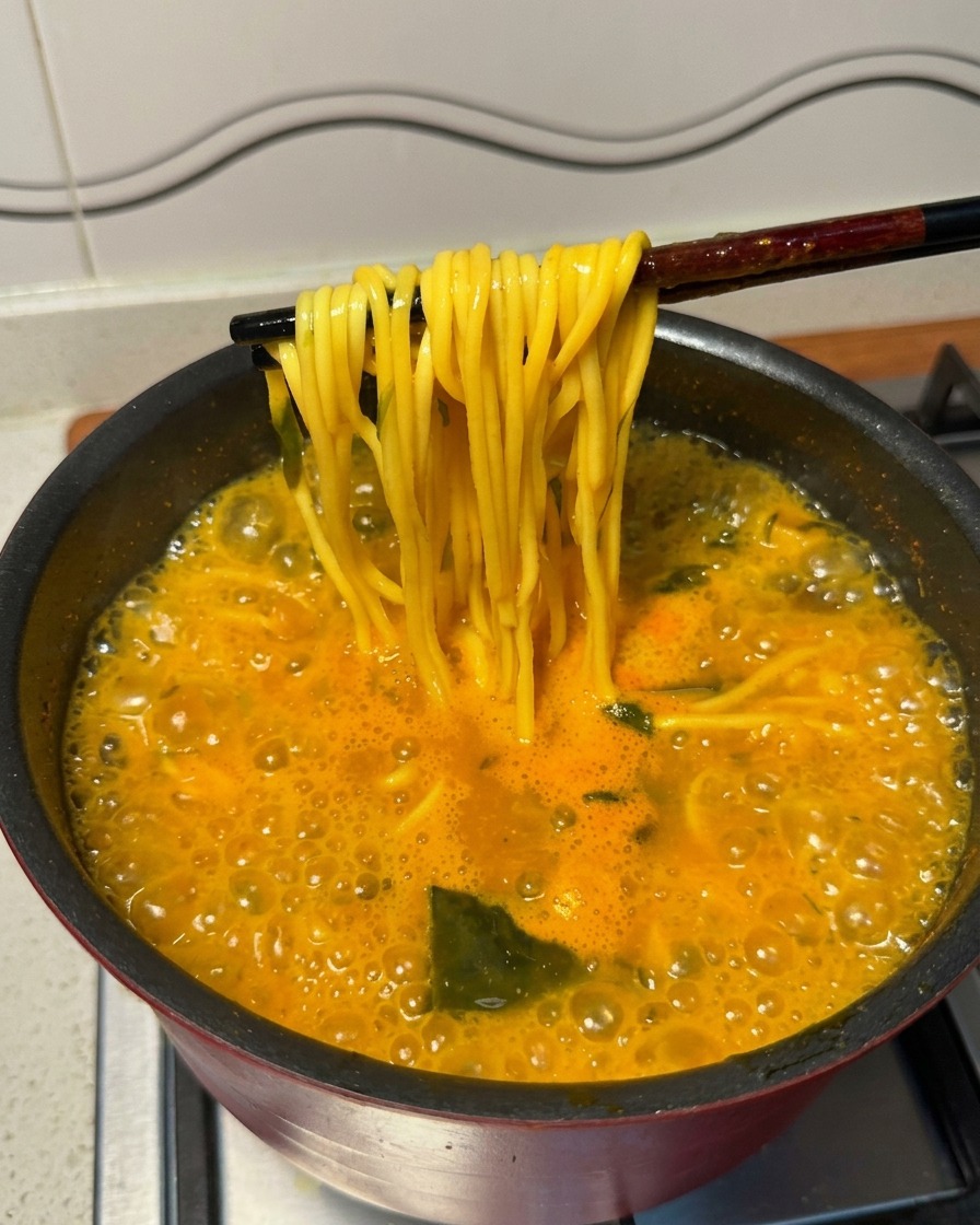 Black chopsticks lifting cooked yellow ramen noodles from a vigorously boiling pot of spicy soup.