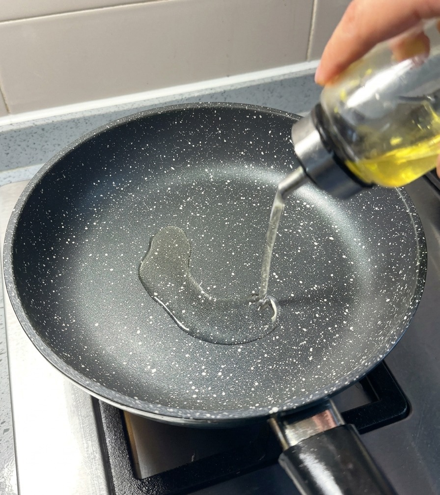 Clear cooking oil being poured from a glass dispenser into a heated, black speckled frying pan.