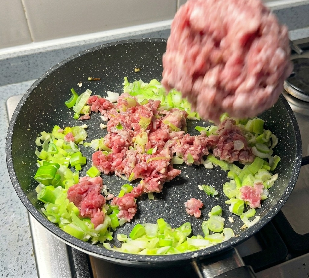 A portion of raw minced pork being dropped into a pan with sautéed green onions.