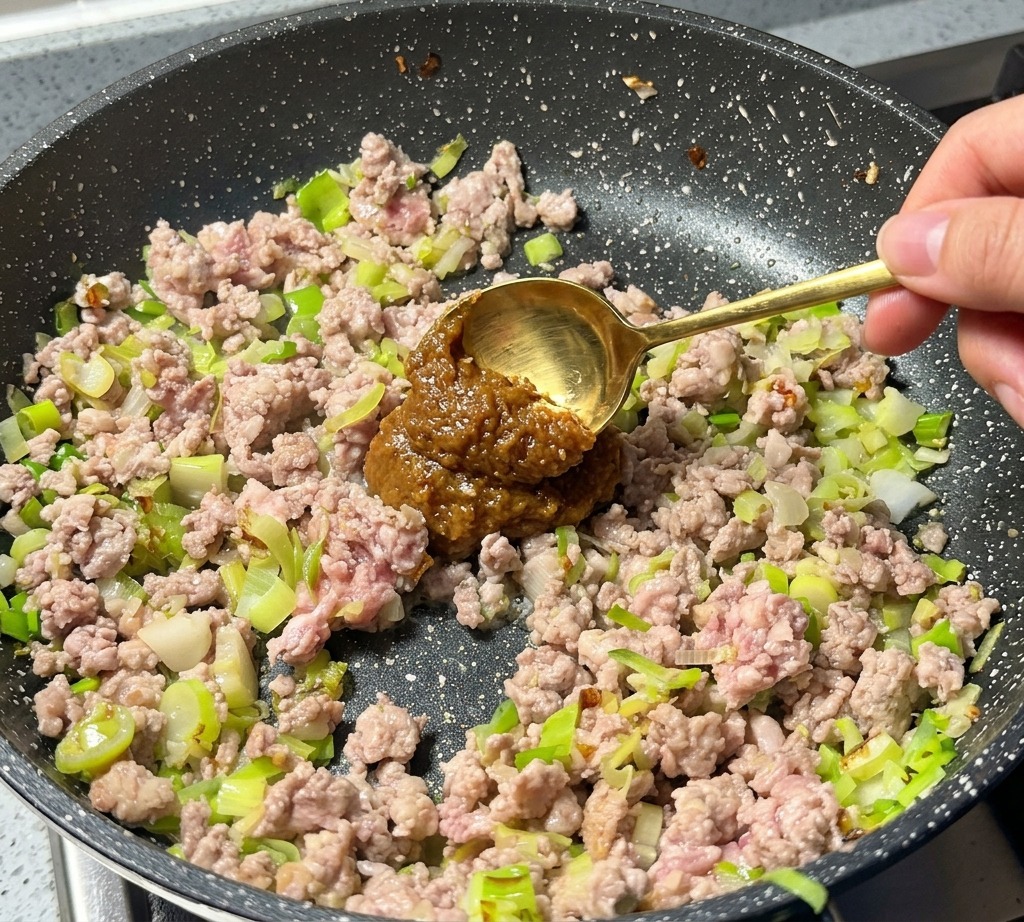 A golden spoon adding a dollop of brown miso paste to browned minced meat and green onions in a pan.