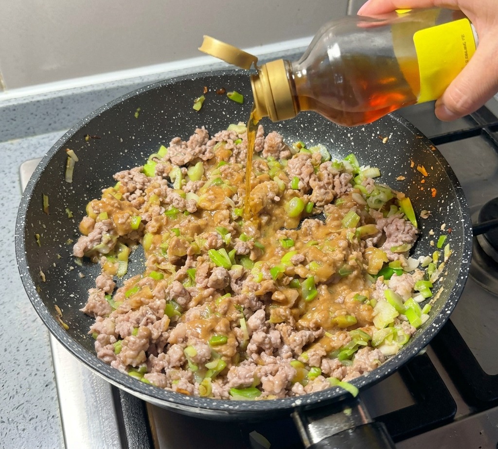 Sesame oil being poured from a bottle over a mixture of minced meat, green onions, and miso in a speckled pan.