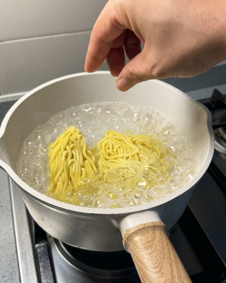 A hand dropping two portions of raw yellow ramen noodles into a pot of rapidly boiling water.