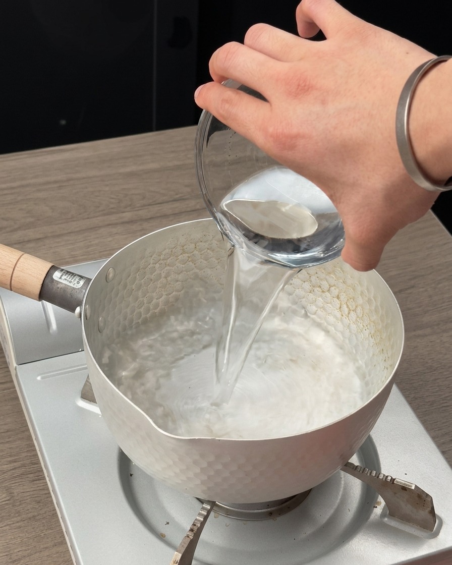 Clear water being poured from a glass measuring bowl into a small pot on a portable gas stove.