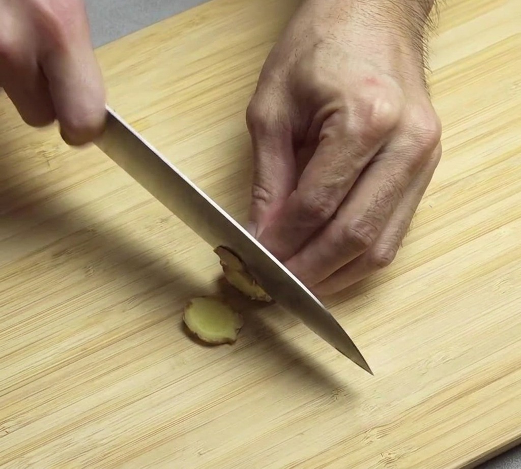 Hands holding a chefs knife, actively slicing a piece of fresh ginger root on a wooden bamboo cutting board.