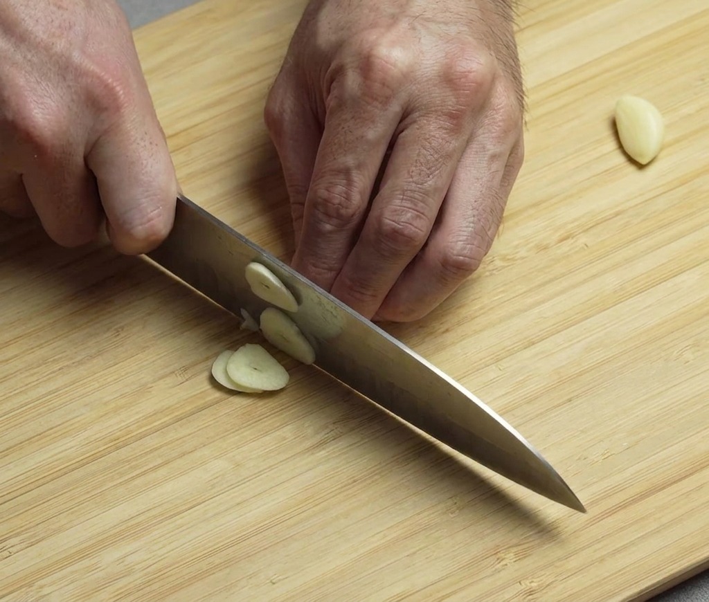 Hands using a knife to finely slice and mince peeled garlic cloves on a wooden cutting board.
