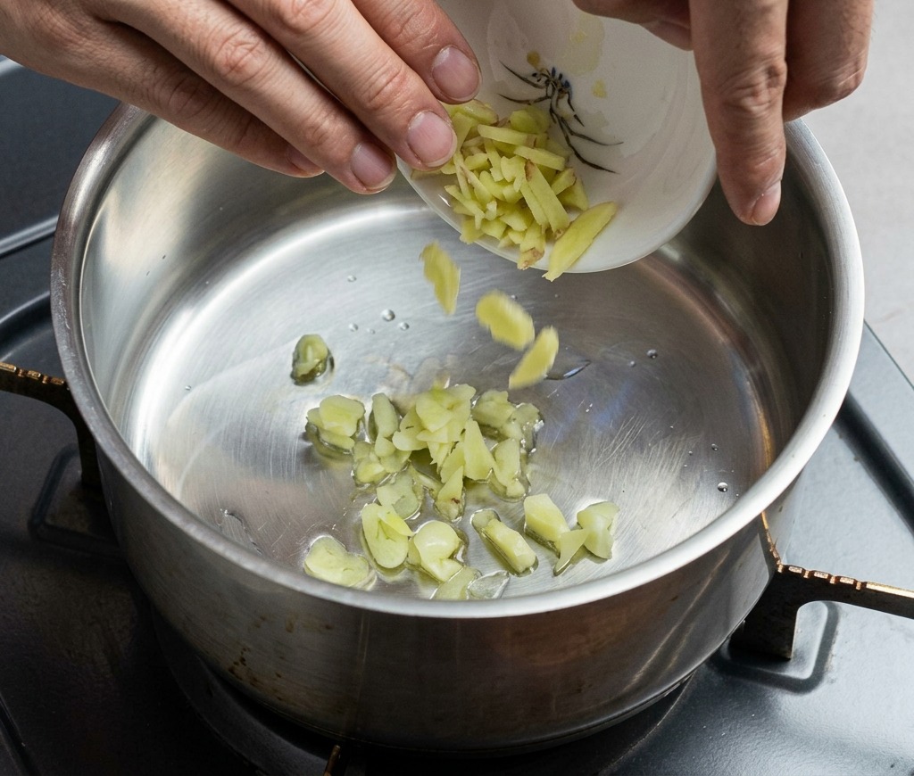 Hands carefully pouring chopped ginger and garlic from a small decorative bowl into a hot stainless steel pot.