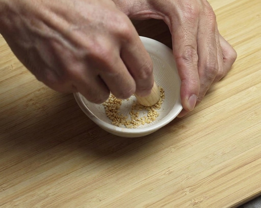 Two hands using a small wooden pestle to crush white sesame seeds inside a white grooved bowl.