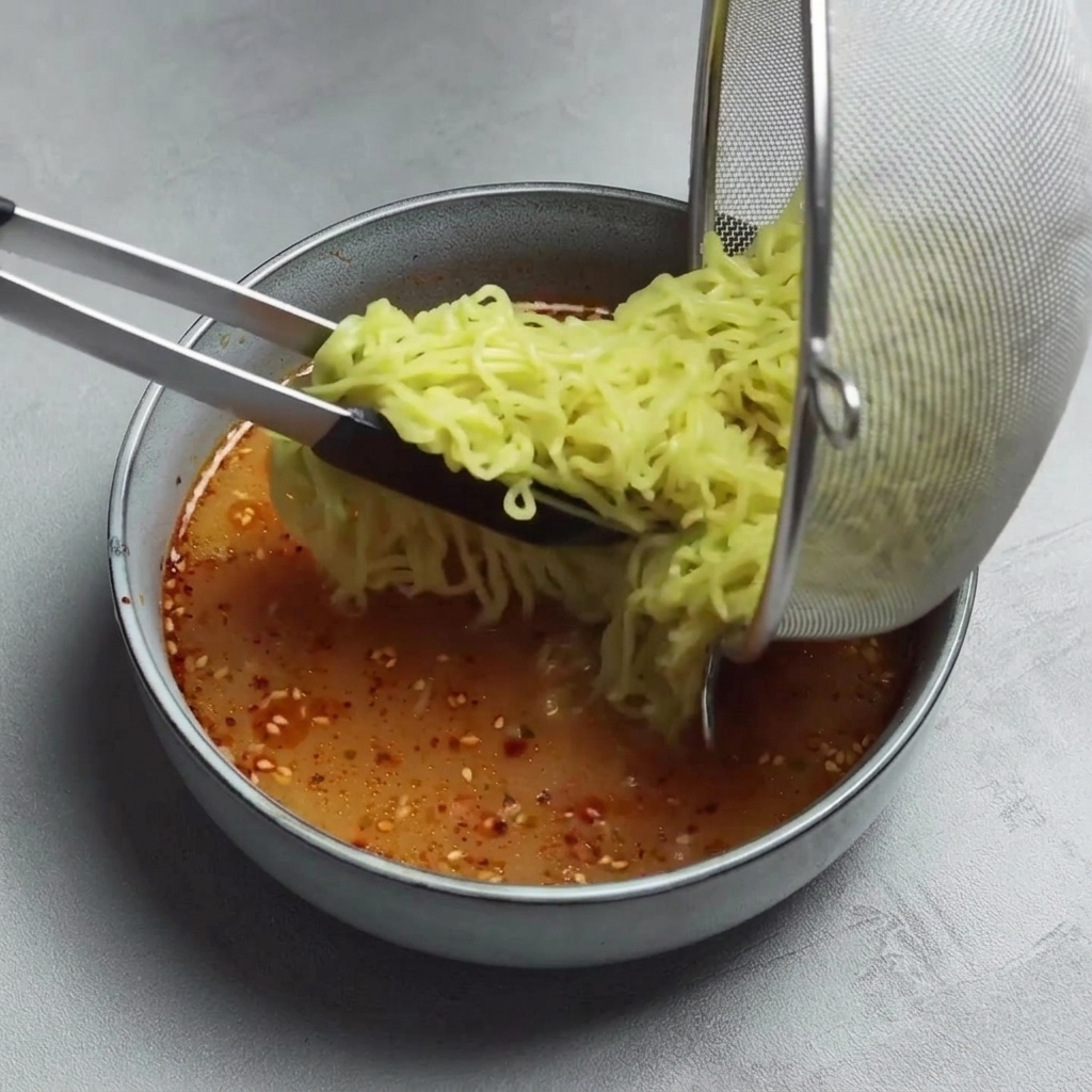 Tongs transferring cooked, yellow ramen noodles from a metal mesh strainer into a bowl of spicy red broth.