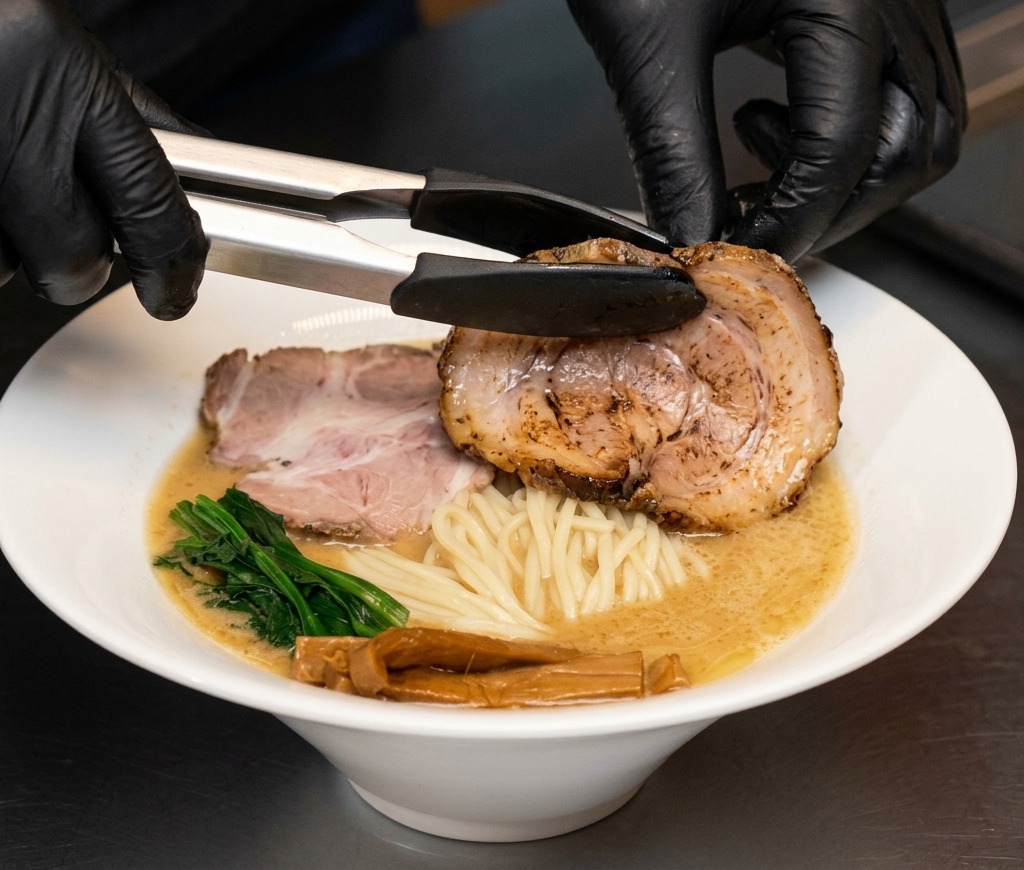 Tongs placing a large slice of chashu pork belly into a bowl of ramen.