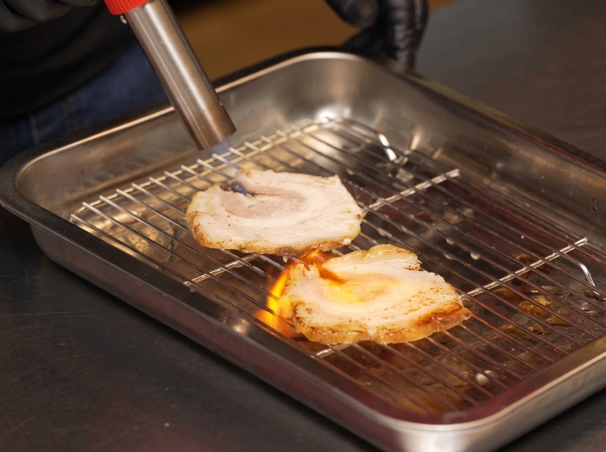 A chef using a blowtorch to sear slices of chashu pork on a metal wire rack.