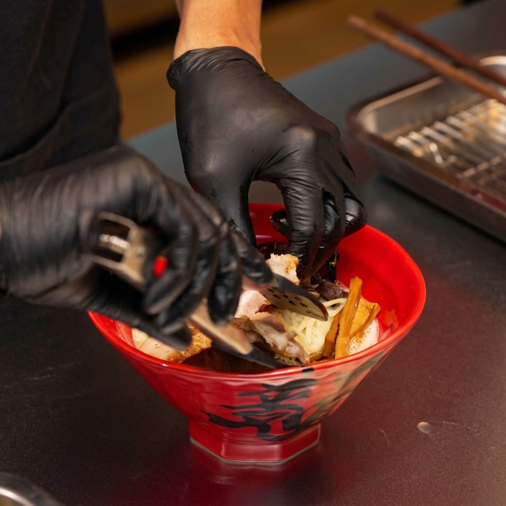 Gloved hands using tongs to place seared chashu pork into a red bowl of miso ramen.