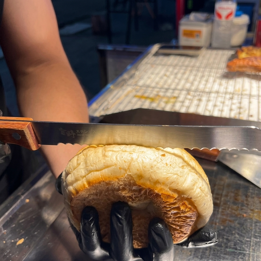 A hand using a serrated knife to gently cut a sesame seed hamburger bun in half horizontally.