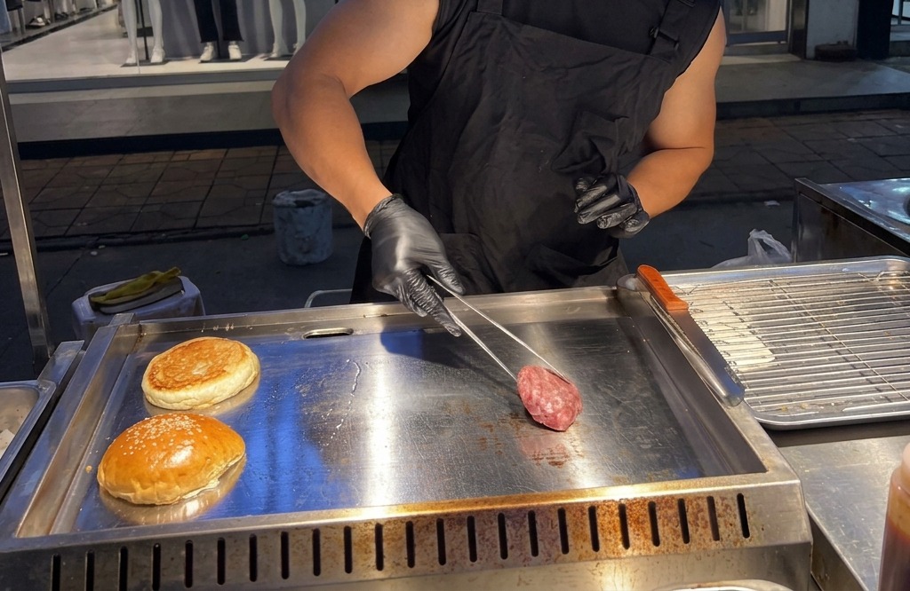 A person placing a round ball of raw ground beef onto a smoking hot metal griddle using metal tongs.